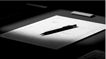A monochrome drafting notebook with a pen laid across gridded paper.